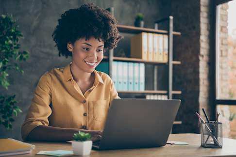 A young woman with curly hair, wearing a mustard yellow shirt, is sitting at a wooden desk, smiling while working on a laptop. The background includes a stylish gray wall with a modern bookshelf filled with colorful binders. Natural light streams in from a large window, creating a bright and inviting workspace atmosphere. A small potted plant and stationery holder with pens and pencils are neatly placed on the desk.
