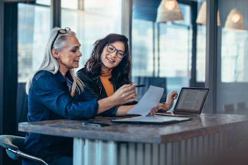 Two women engaged in a professional discussion sit at a polished wooden table in a modern office setting. One woman, with long gray hair in a denim jacket, points at a document she's holding, while the other, wearing glasses and a black leather jacket over a mustard top, listens attentively. A laptop displaying text is open in front of them, suggesting a collaborative work environment. The background features contemporary decor with pendant lights and large windows, creating a bright and focused atmosphere.
