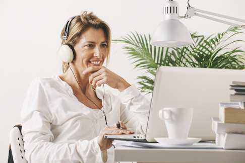 A woman is sitting at a desk in a modern workspace, wearing large headphones and looking at a laptop with a smile. She is dressed in a white blouse, and the setting includes a white table lamp, a stack of books, and a cup on a saucer. A green potted plant adds a touch of nature in the background. The overall mood is professional and focused, with a sense of comfort and engagement.