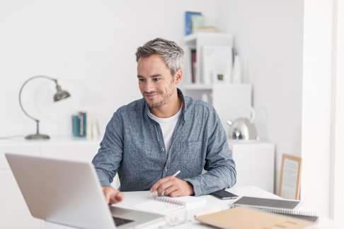 A man with salt-and-pepper hair sits at a white desk in a bright, airy office. He is wearing a casual blue shirt and has a slight smile while working on a laptop. On the desk, there are a notebook, a pen, and a smartphone. In the background, there are white shelves with books and various decorative items, creating a clean and modern atmosphere.