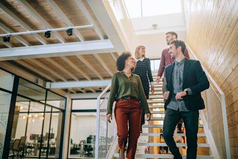 A diverse group of four young professionals, two women and two men, walking down a modern office staircase. The office is bright, with large windows and wooden paneling on the walls, creating a warm and inviting atmosphere. The individuals are engaged in conversation, suggesting a collaborative and friendly work environment. The style is contemporary, emphasizing open spaces and modern architecture.