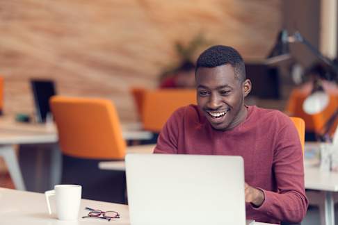 The image shows a young man sitting at a desk in a modern office environment, engaging with his laptop while smiling. He is wearing a red sweater and appears to be focused and pleased with what he is seeing on the screen. The office features a contemporary design with bright colors, including orange chairs and a blurred wall backdrop, contributing to a lively and positive atmosphere. A white coffee mug and a pair of glasses rest on the desk, adding to the workspace setting.