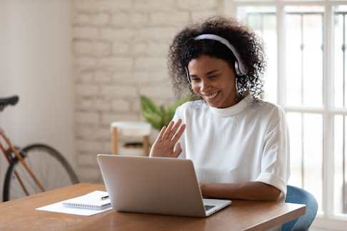 A young woman with curly hair sits at a wooden desk, engaging in a video call on a laptop. She is wearing a white shirt and headphones, waving and smiling at the screen. The setting is a bright, modern room with a brick wall, a bicycle, and plants in the background, creating a friendly and professional atmosphere.